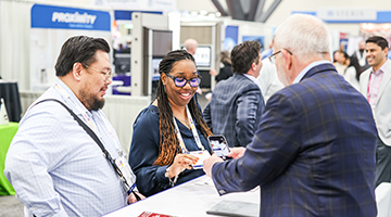 two attendees standing at table talking with exhibitor at booth